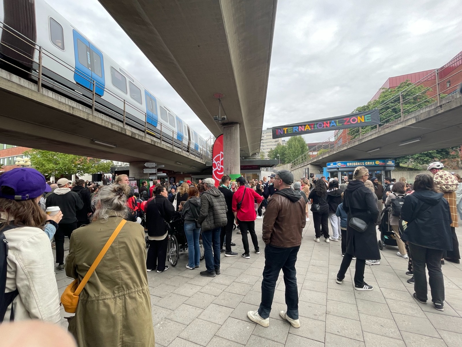 Folksamling under bron vid Bredängs torg med tunnelbanan som passerar förbi och en handsydd INTERNATIONAL ZONE-banner.