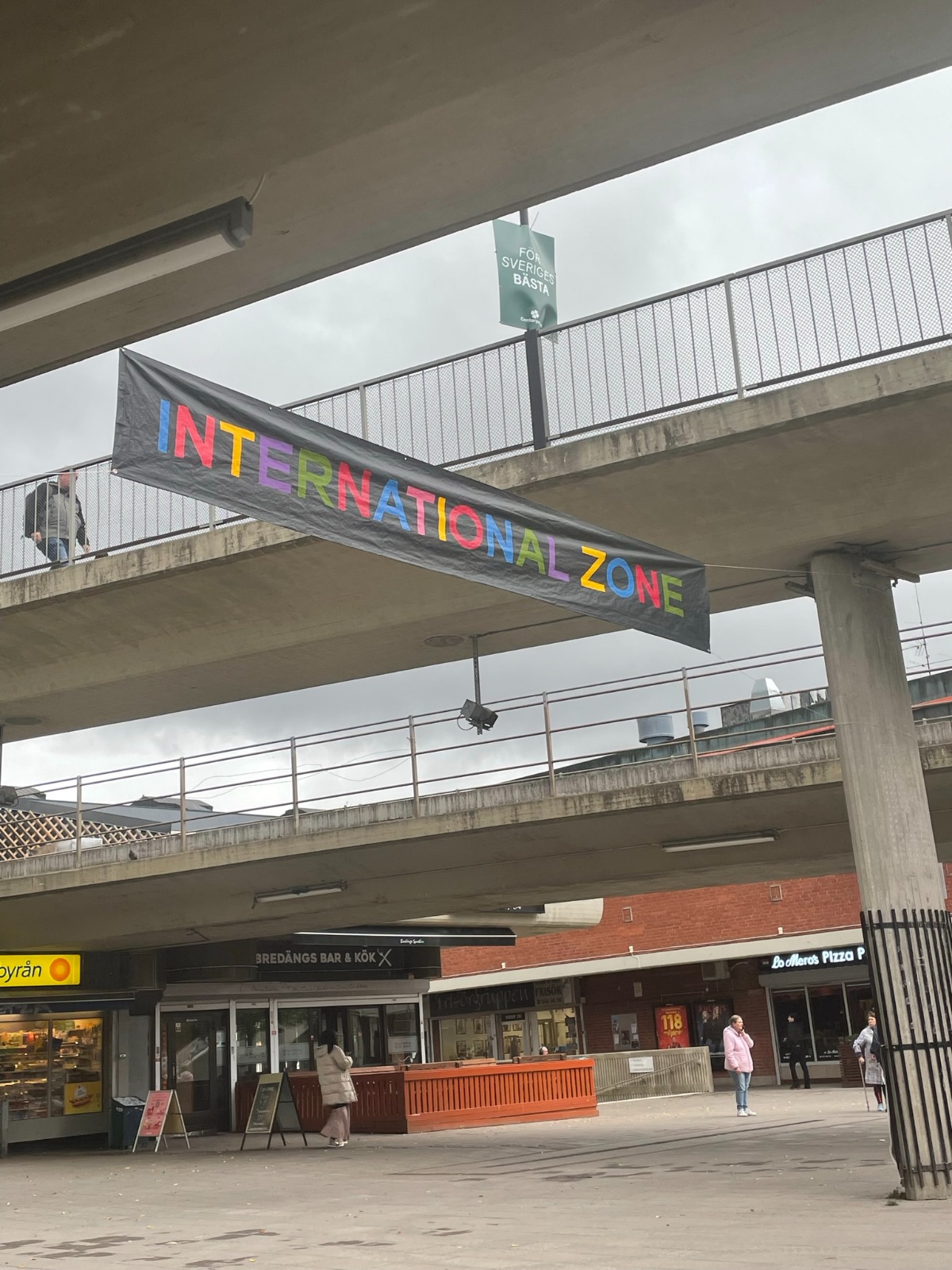 Den färgglada INTERNATIONAL ZONE-bannern spänd mellan bron och räcket en mulen dag.
